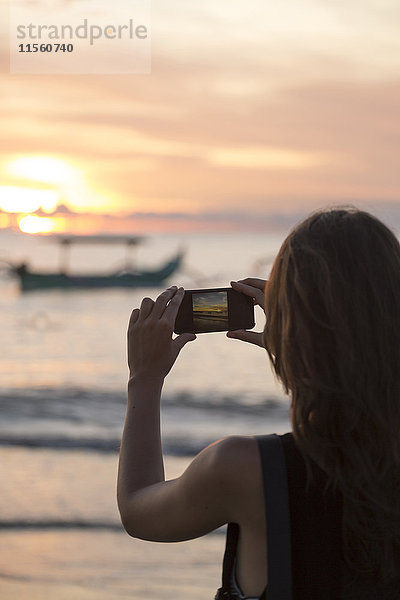 Indonesien  Bali  Frau beim Fotografieren des Sonnenuntergangs über dem Meer