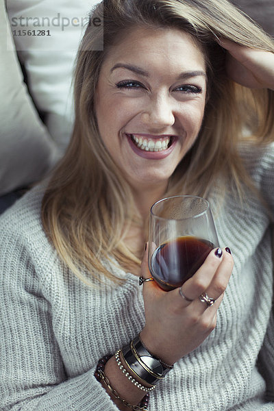 Porträt einer lachenden blonden Frau mit einem Glas Kaffee auf der Couch zu Hause.