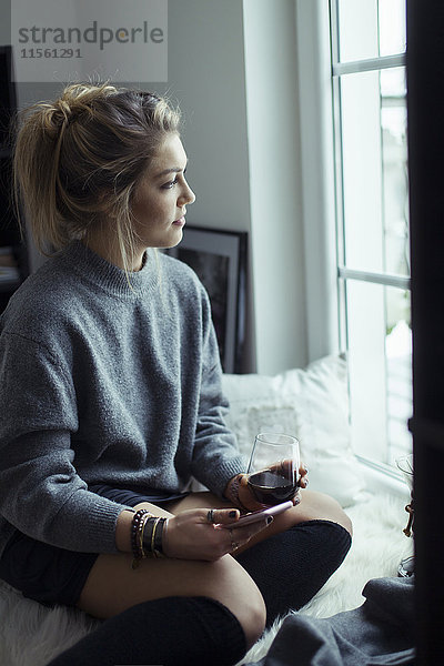 Junge Frau mit Tasse Kaffee und Smartphone durchs Fenster schauend