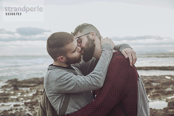 Junges schwules Paar  das sich vor dem Meer umarmt und küsst.