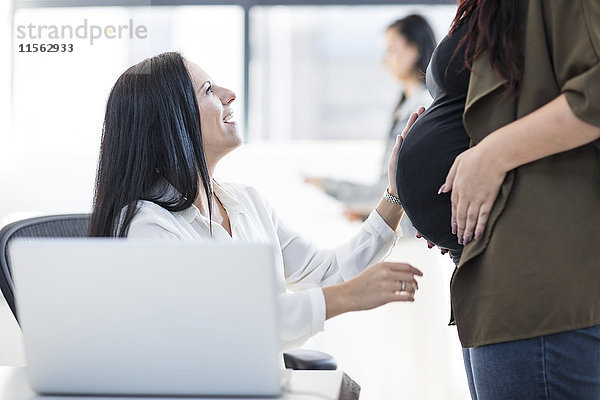 Frau im Büro berührt einen schwangeren Kollegen Bauch