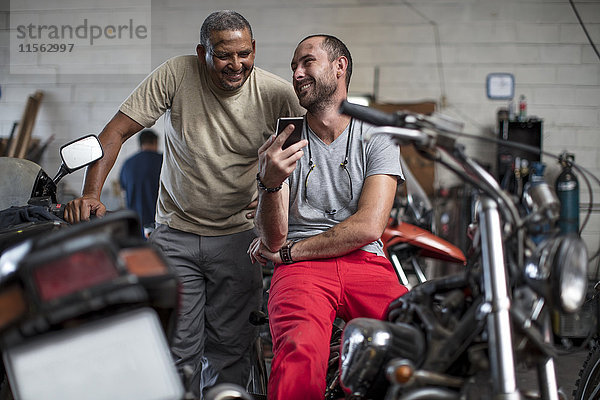 Zwei lächelnde Mechaniker in der Motorradwerkstatt beim Blick aufs Handy
