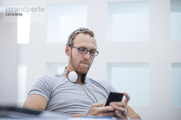 Junger Mann mit Kopfhörern um den Hals  der Textnachrichten auf seinem Telefon liest.
