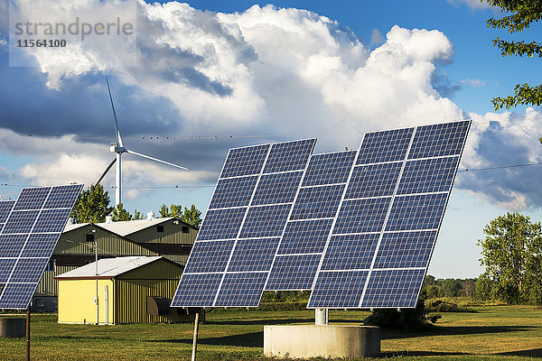 Große Sonnenkollektoren auf einem Bauernhof mit Metallscheunen und einer großen Metallwindmühle im Hintergrund mit Wolken und blauem Himmel; Ontario  Kanada'.