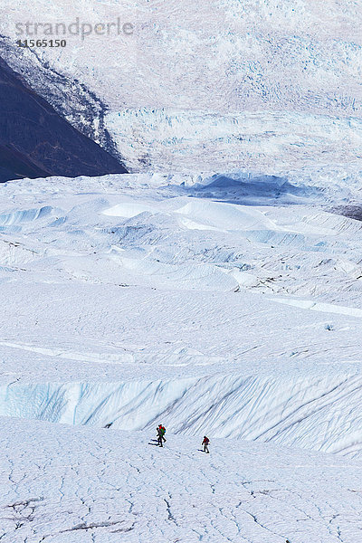 Menschen überqueren den Root-Gletscher im Wrangell-St. Elias-Nationalpark  um an einer geführten Eisklettertour teilzunehmen. Im Hintergrund ist der Stairway-Eisfall zu sehen  Southcentral Alaska  USA