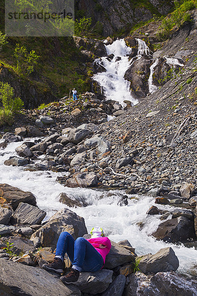 Eine Frau macht eine Pause vom Seekajakfahren im Kenai Fjords National Park und entspannt sich an einem rauschenden Bach  während ihr Partner einen Wasserfall erkundet; Alaska  Vereinigte Staaten von Amerika'.