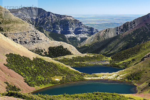 Zwei farbenfrohe alpine Bergseen zwischen felsigen und bewaldeten Berghängen mit blauem Himmel und Prärie in der Ferne; Waterton  Alberta  Kanada'.