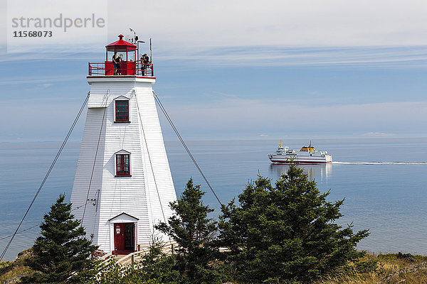 Weißer Leuchtturm mit Menschen auf der obersten Plattform mit Blick auf die Fährüberfahrt in der Ferne und den dunstigen blauen Himmel; North Head  New Brunswick  Kanada'.