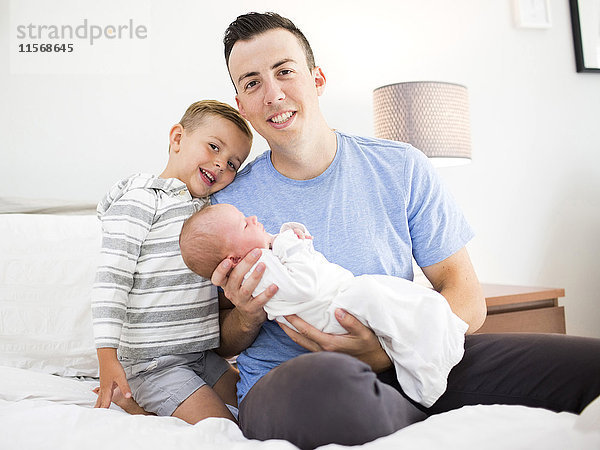 Vater trägt Tochter (0-1 Monate) und sitzt mit Sohn (4-5) auf dem Bett