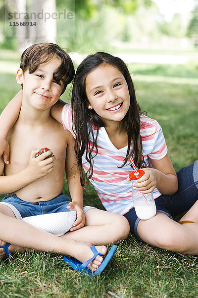 Junge (6-7) und Mädchen (10-11) sitzen im Sommer im Gras