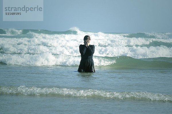 Frau in schwarzem Kleid mit Maske  stehend in der Brandung im Meer