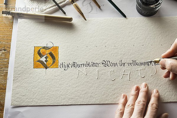 Kalligraphie-Atelier  Hand schreibt Text mit Feder und Stift  Buchstabe S mit Gold beschichtet auf Torchonpapier  Handprägung sichtbar  Tintenfass und Federhalter auf der Rückseite  Seebruck  Oberbayern  Deutschland  Europa