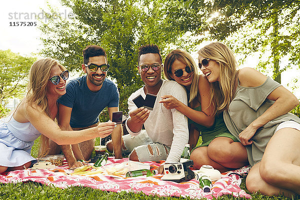 Fünf erwachsene Freunde schauen sich beim Picknick im Park einen Film von einer Sofortbildkamera an