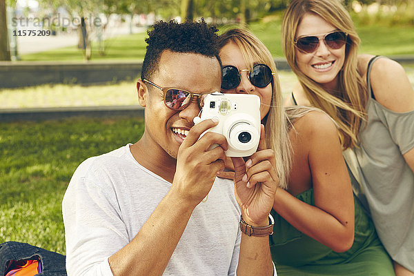 Junger Mann und Freunde fotografieren mit Sofortbildkamera im Park