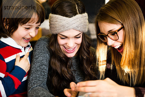 Zwei junge Frauen und ein Junge schauen und lachen über ein Smartphone im Cafe