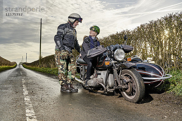 Älterer männlicher Motorradfahrer im Gespräch mit seinem Enkel  der auf einem Motorrad am ländlichen Straßenrand sitzt