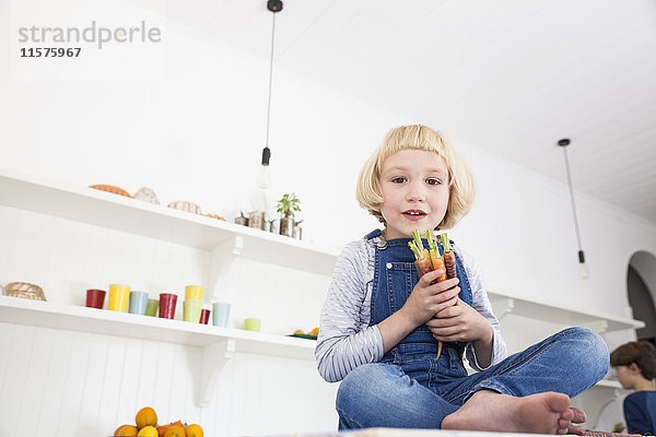 Porträt eines süßen Mädchens  das auf dem Küchentisch sitzt und einen Strauß bunter Karotten hält