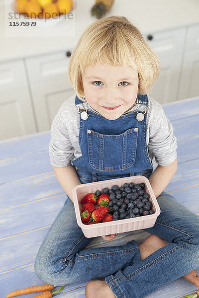 Porträt eines süßen Mädchens  das auf der Küchentheke ein Kännchen mit Blaubeeren und Erdbeeren hält