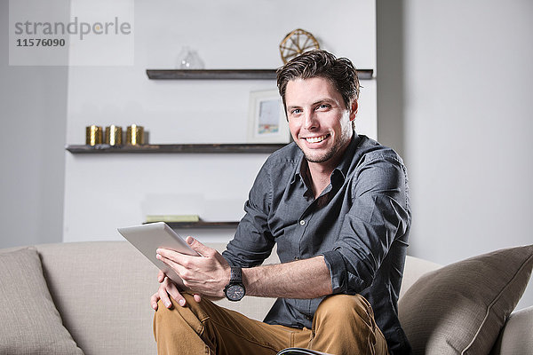 Porträt eines jungen Mannes  der auf einem Sofa sitzt  ein digitales Tablet hält und lächelt