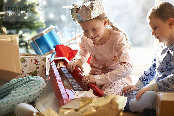 Mädchen und Bruder auf dem Wohnzimmerboden mit Blick auf ein Weihnachtsgeschenk für eine Spielzeuggitarre