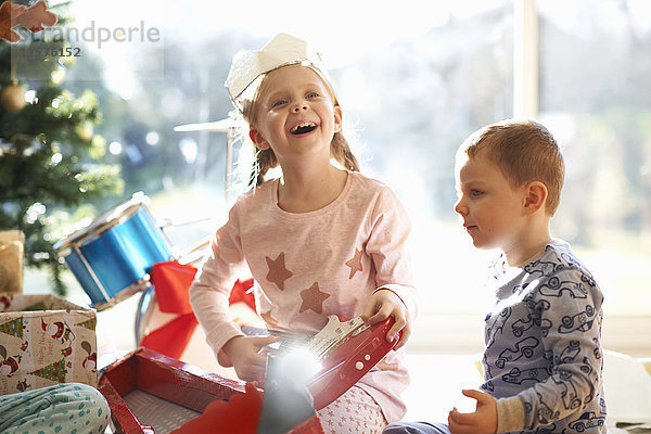 Aufgeregtes Mädchen und Bruder auf Wohnzimmerboden mit Spielzeuggitarre als Weihnachtsgeschenk