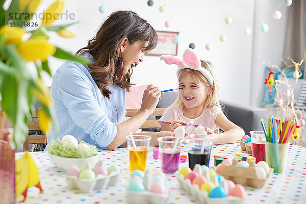 Frau malt der Tochter die Nase  während sie bei Tisch Ostereier malt
