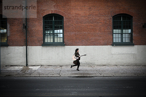 Junge Frau rennt mit Akustikgitarre auf dem Bürgersteig der Stadt
