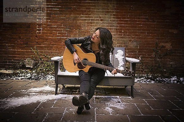 Junge Frau auf verschneitem Bürgersteigsitz in der Stadt spielt Akustikgitarre