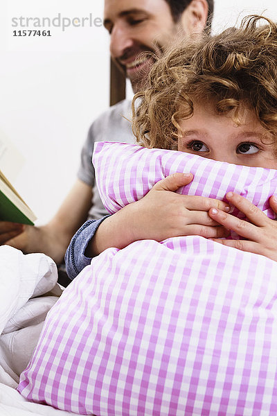 Mädchen umarmt Kissen  während der Vater im Bett ein Märchenbuch liest