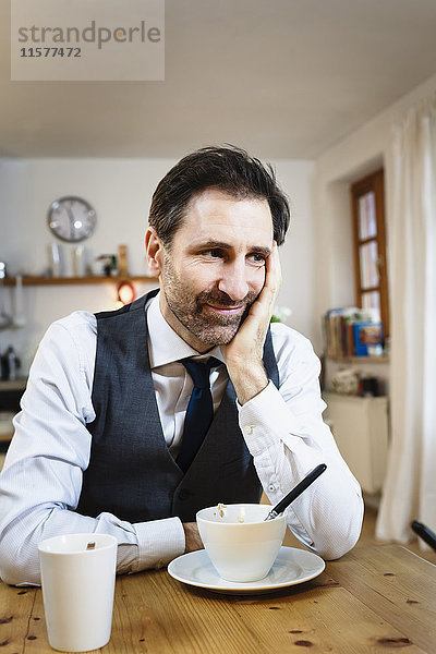 Lächelnder reifer Mann mit Kinn zur Hand beim Frühstück in der Küche