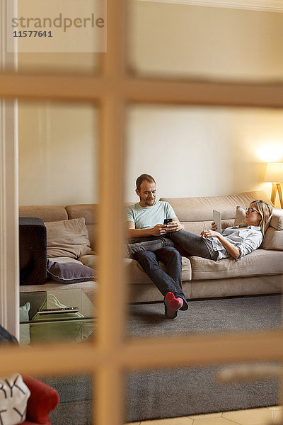 Mittleres erwachsenes Paar entspannt auf dem Sofa  Mann benutzt Smartphone  Frau benutzt digitales Tablet