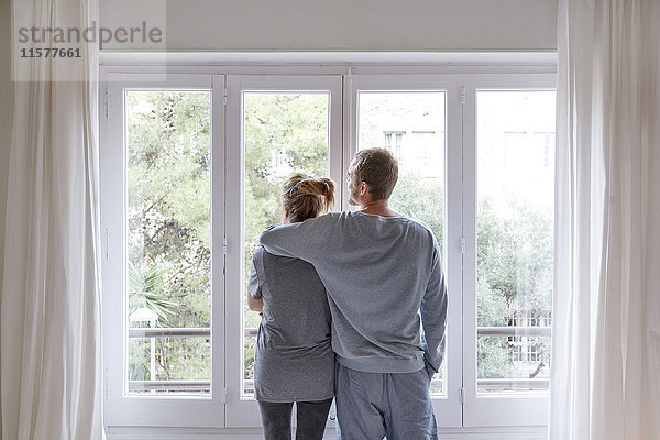Mittleres erwachsenes Paar zu Hause  Blick aus dem Fenster  Mann umarmt Frau  Rückansicht