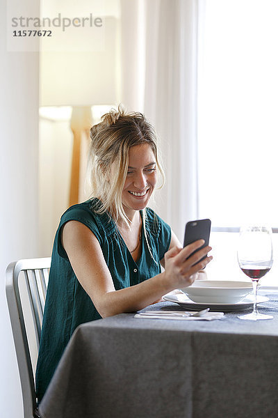 Frau sitzt am Esstisch  benutzt Smartphone und lächelt
