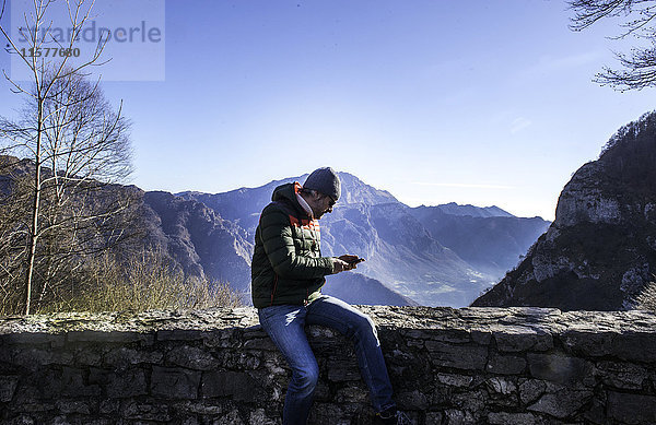 Mann sitzt auf einer Mauer in ländlicher Umgebung und schaut auf ein Smartphone  Italien