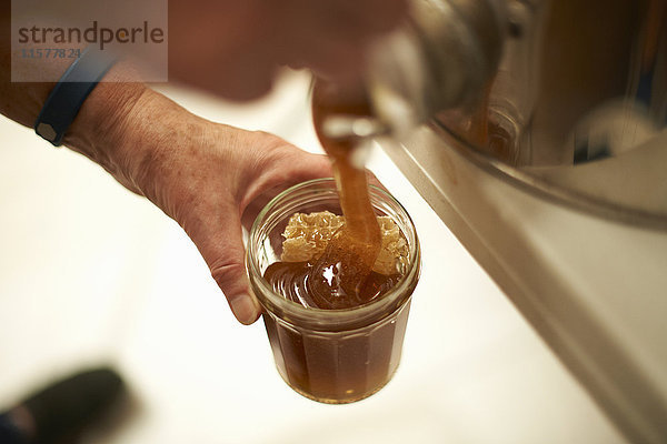 Hände einer Imkerin gießen Honig aus Küchenbottich in ein Glas