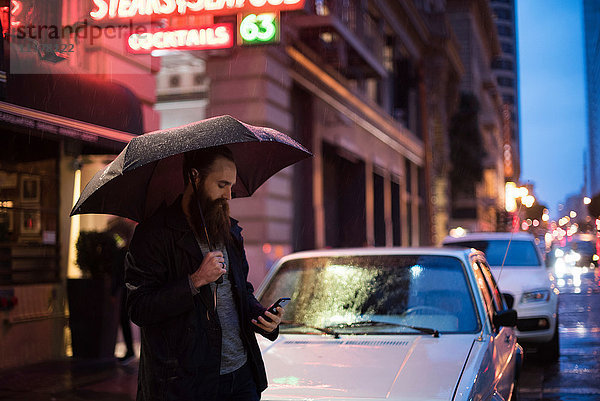 Mittelgroßer erwachsener Mann  der nachts in der Stadt spazieren geht  einen Regenschirm benutzt und auf ein Smartphone schaut  Downtown  San Francisco  Kalifornien  USA