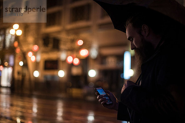 Mittelgroßer erwachsener Mann  der nachts in der Stadt spazieren geht  einen Regenschirm benutzt und auf ein Smartphone schaut  Downtown  San Francisco  Kalifornien  USA