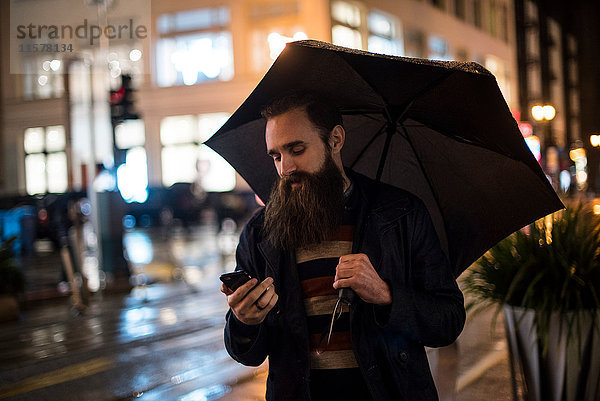 Mittelgroßer erwachsener Mann  der nachts in der Stadt spazieren geht  einen Regenschirm benutzt und auf ein Smartphone schaut  Downtown  San Francisco  Kalifornien  USA