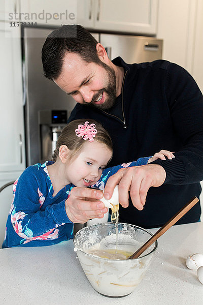 Mittelgroßer erwachsener Mann und Tochter schlagen zusammen ein Ei in eine Rührschüssel an der Küchentheke
