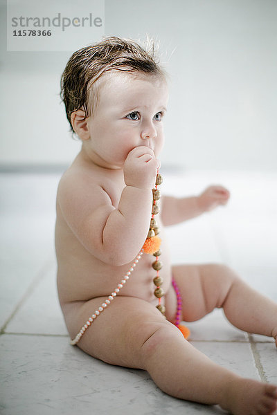Nacktes weibliches Kleinkind sitzt auf Badezimmerboden und probiert Perlen
