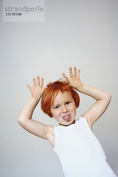 Porträt eines rothaarigen Mädchens im Haus  das seine Zunge herausstreckt und seine Hände in die Luft legt.