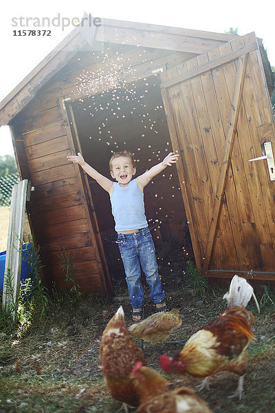 Kleiner Junge wirft fröhlich Samen zu den Hühnern vor einem Hühnerstall.