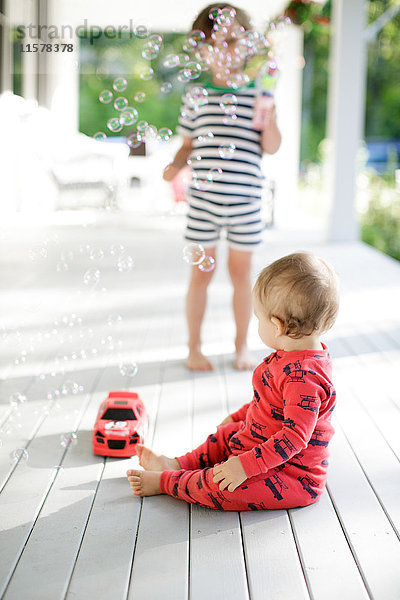 Männliches Kleinkind sitzt auf der Veranda und beobachtet den Jungen  wie er Blasen bläst.