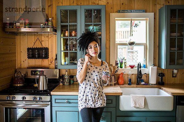 Porträt einer jungen Frau  die in der Küche steht und ein heißes Getränk hält.