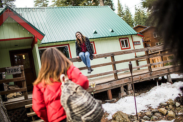 Mädchen sitzt auf dem Zaun der Hütte  ihre Freundin geht auf sie zu.