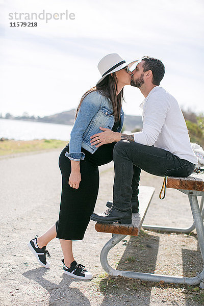 Schwangeres reifes Paar küsst sich auf Parkbank an der Küste