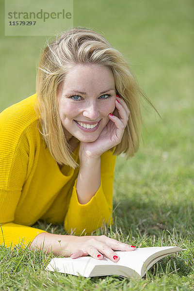 Blonde hübsche Frau beim Lesen eines Buches im Park lächelnd vor der Kamera