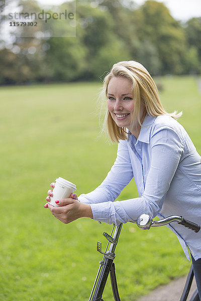 Hübsche blonde Frau mit Fahrrad und Kaffee im Park
