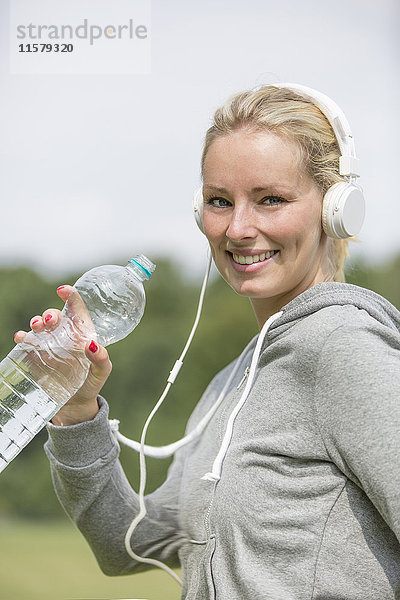 Hübsche Frau macht Sport im Park  trinkt Wasser und lächelt in die Kamera.