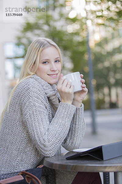 Hübsche blonde Frau genießt eine Tasse Kaffee in der Innenstadt und lächelt vor der Kamera.